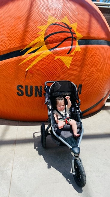 A baby sits in a stroller in front of a giant basketball. Phoenix Suns logo is painted on the basketball.