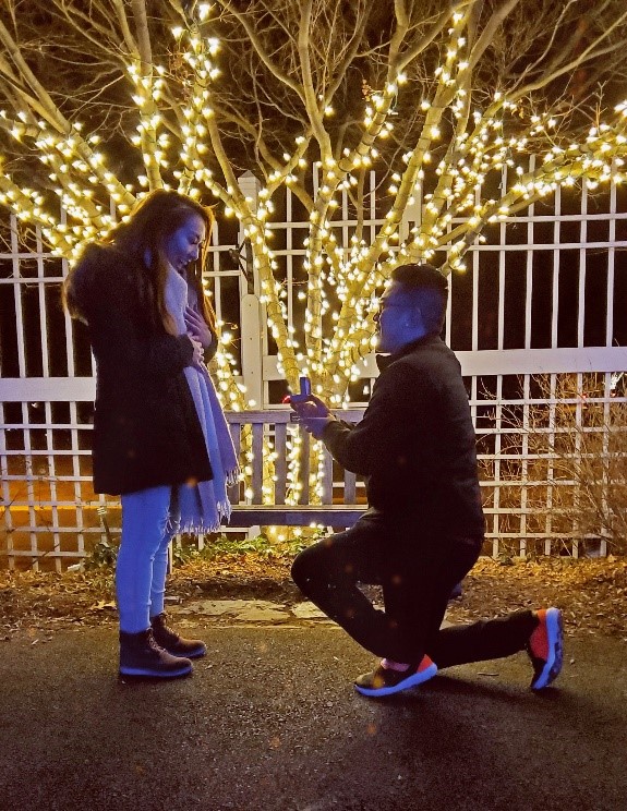 A man rests on one knee and proposes to a woman in front of Christmas lights at night