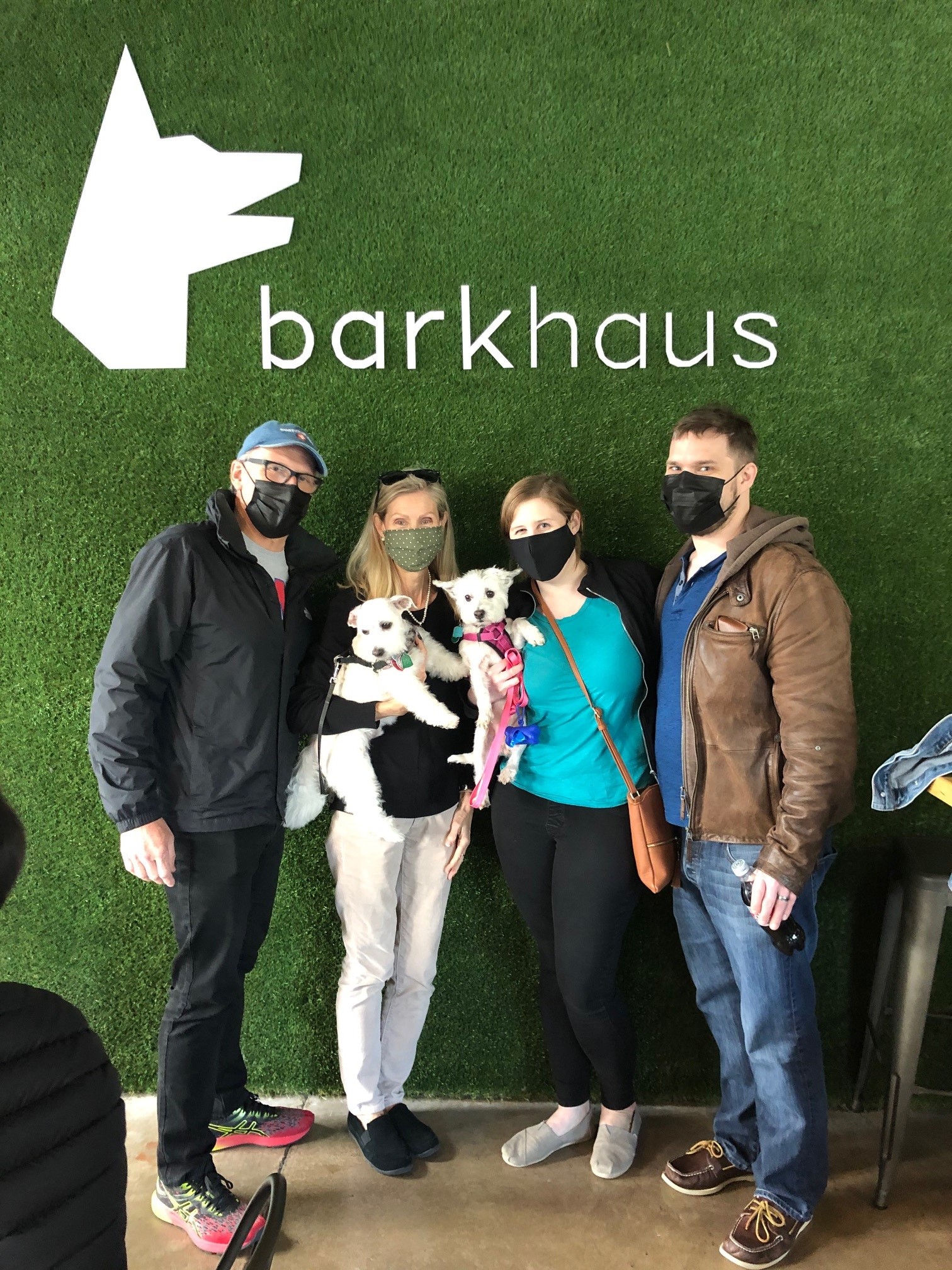 Four masked adults and two dogs take a photo in front of a green backdrop. Text in the background reads Barkhaus with a dog logo.