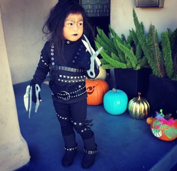 Young child in all-black attire with metal-studded belts across the entire body, black lipstick, and plastic mini blades representing scissors on her hands.