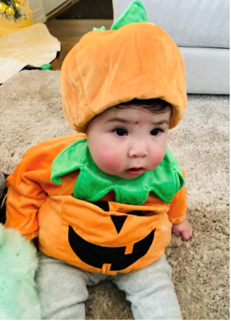 Infant dressed in a round orange costume with 2 black circles, one triangle, and a broken smile on the abdomen. The infant wears a green collar with an orange and green hat.