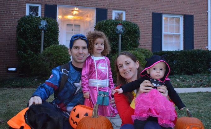 From left to right, man dressed in a blue, white, and red long sleeve t-shirt with stars and stripes, little girl in a pink and white dress with silver “S” inside a diamond shape on her chest, woman dressed in red long sleeve shirt with golden cuffs holds a small child in a pink tutu and black long-sleeved hoodie with a bat symbol on her chest. The group of people is surrounded by pumpkins