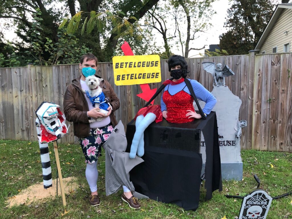 On the left, a man dressed in swimwear with shark costume attached to his leg holds a small white dog wearing a football jersey in his arms. On the right, a woman in a red and blue dress features a black board around her waist to represent a couch with false legs glued on top. They stand in front of decorations made to look like scenes from Beetlejuice the movie. 