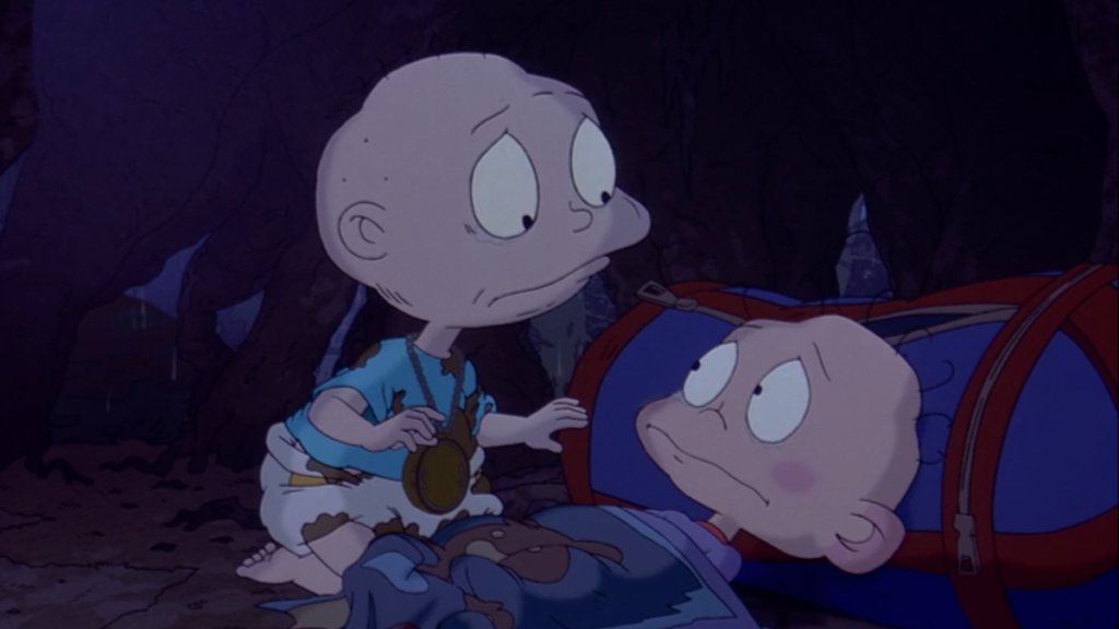 On the left, a bald baby in a white shirt and white and brown diaper looks down at his brother. His brother lays on the ground resting upon a blue and red bag wrapped in a blue blanket.