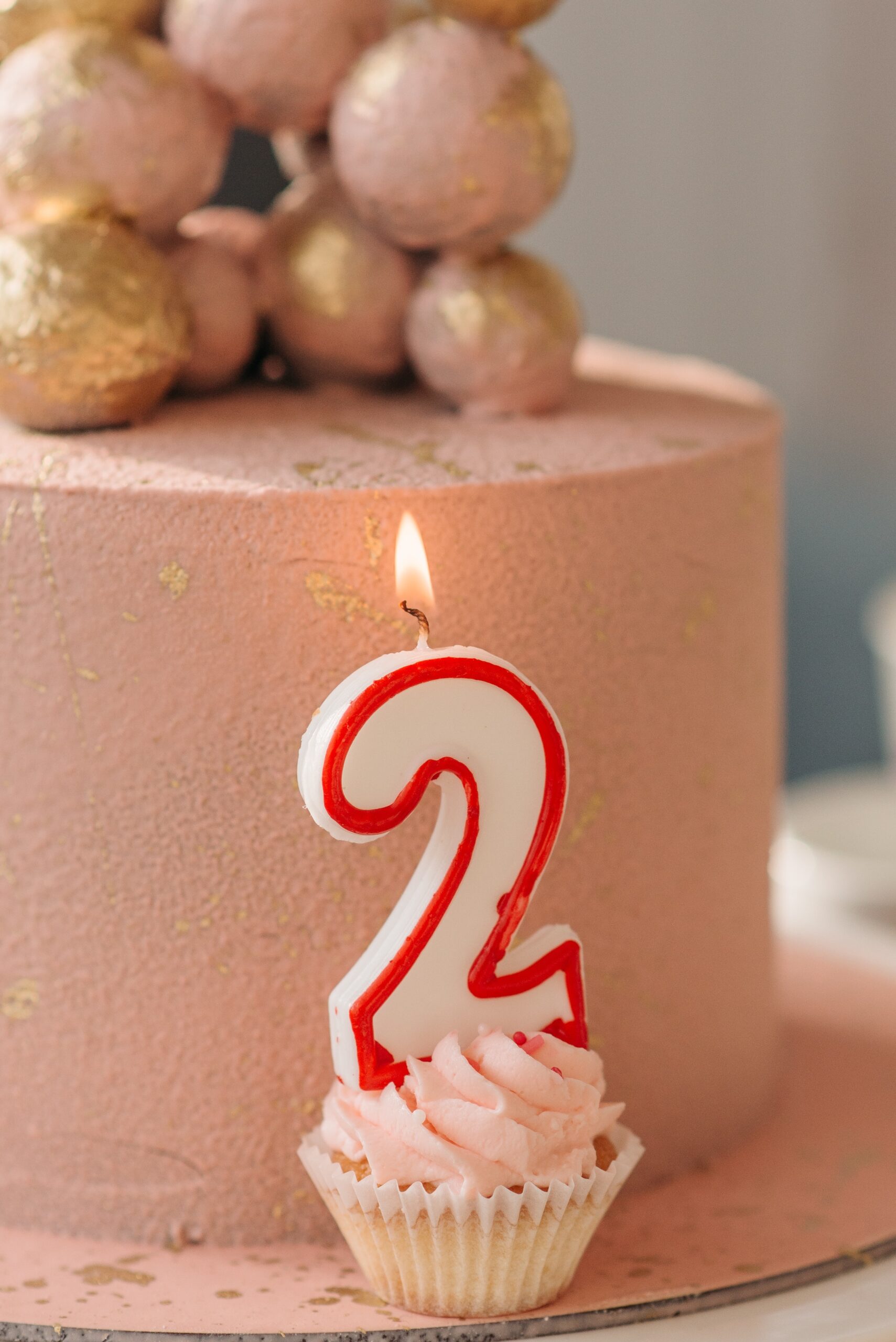 A rose pink cake with gold decorations is in the background. In the foreground, a small cupcake with pink icing sports a lit birthday candle in the shape of the number two.