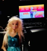 A Barbie doll held in front of a cell phone camera, with rows of theater seats and the screen in the background.