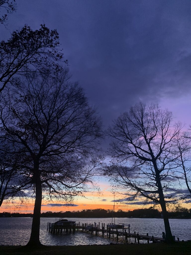 Sunset over a river with a small boat sitting on a dock lift seen from a lawn framed by trees.