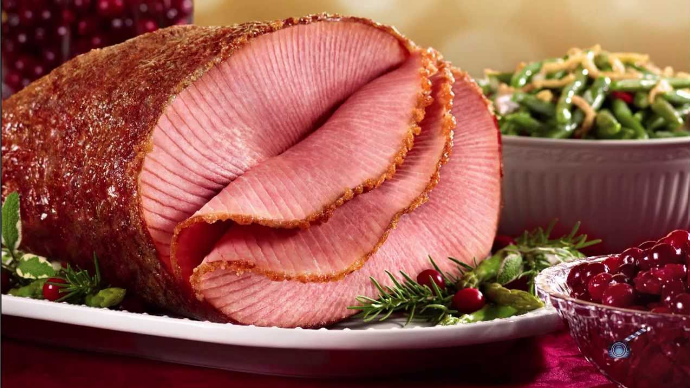 A big baked ham sliced at the front sitting on a decorated tray surrounded by bowls of cranberry sauce and green beans.