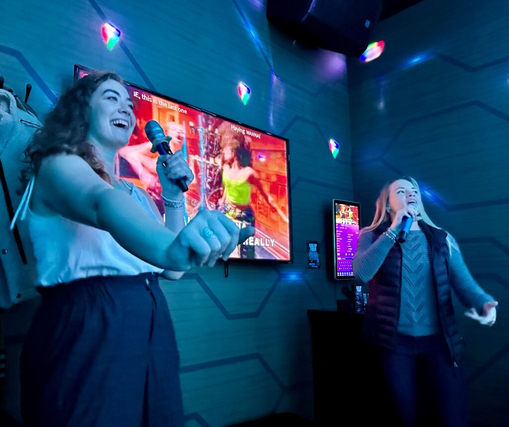 Two people holding microphones sing karaoke in front of a TV screen.